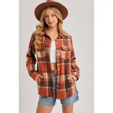 Model wearing a rust and brown plaid flannel shacket with chest pockets, styled with denim shorts and a wide brim hat